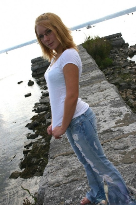Young blonde in ripped jeans takes off her shirt to model in a bra by the sea